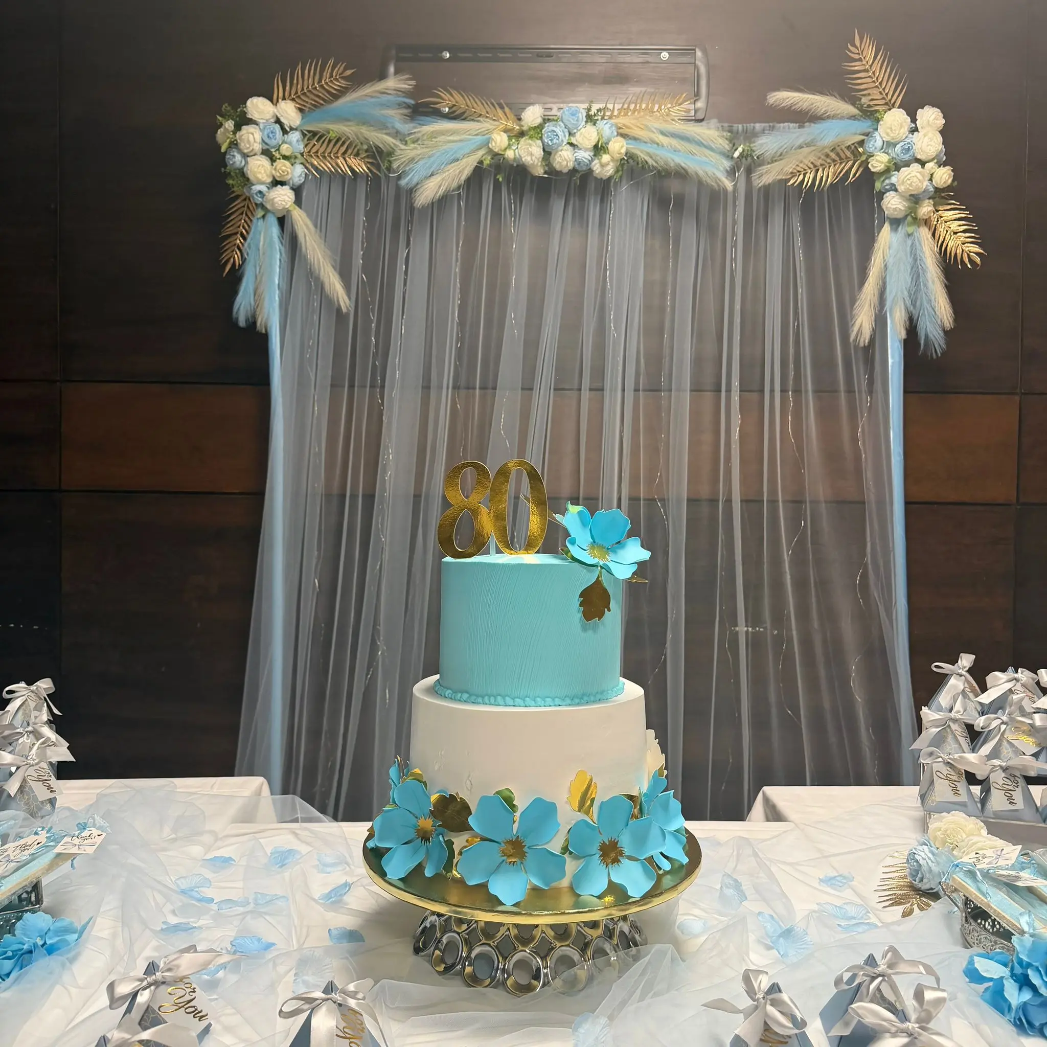 Decoração de mesa de 80.º aniversário com bolo de dois andares azul e branco, flores e cortina de tule iluminada. Ilha do Sal, Cabo Verde.