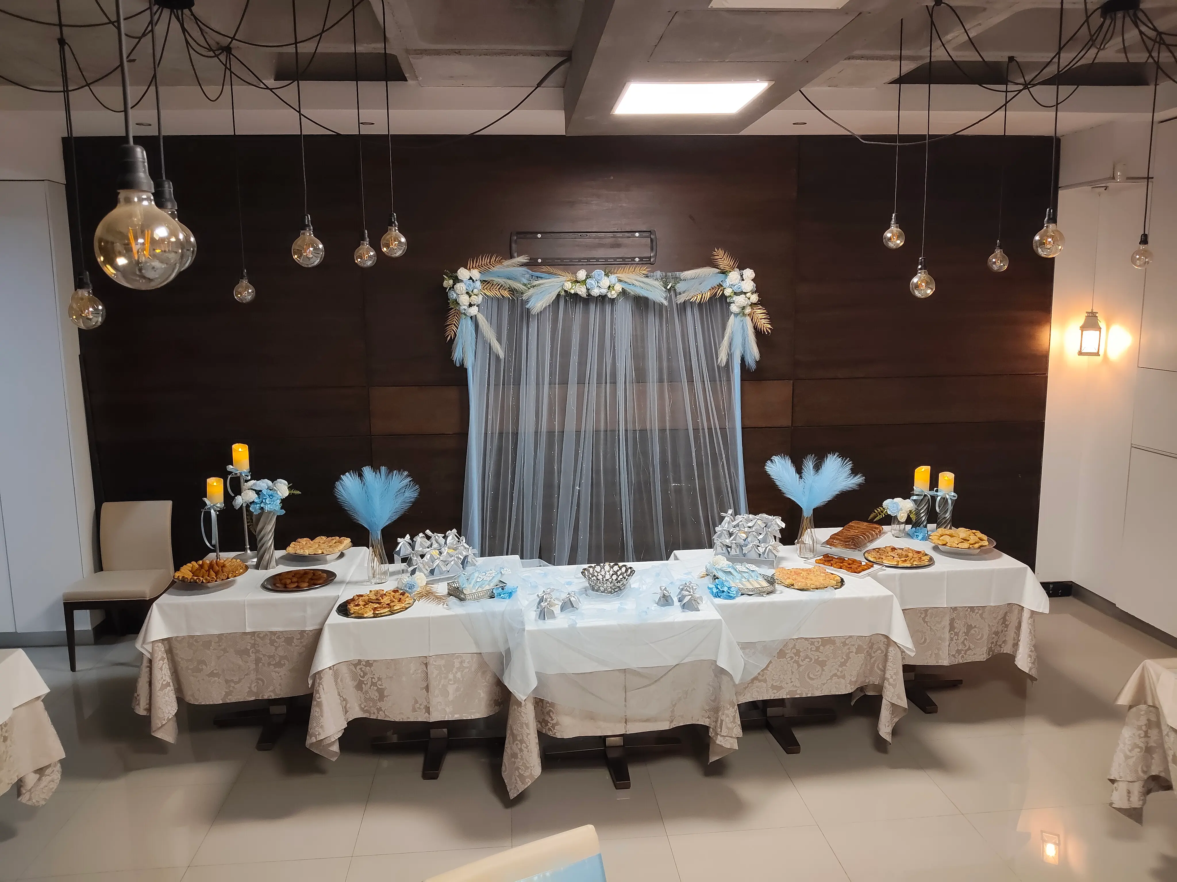 Mesa de aniversário decorada com arranjos florais em tons de azul, doces finos e iluminação suspensa com lâmpadas decorativas. Espargos, Ilha do Sal, Cabo Verde.