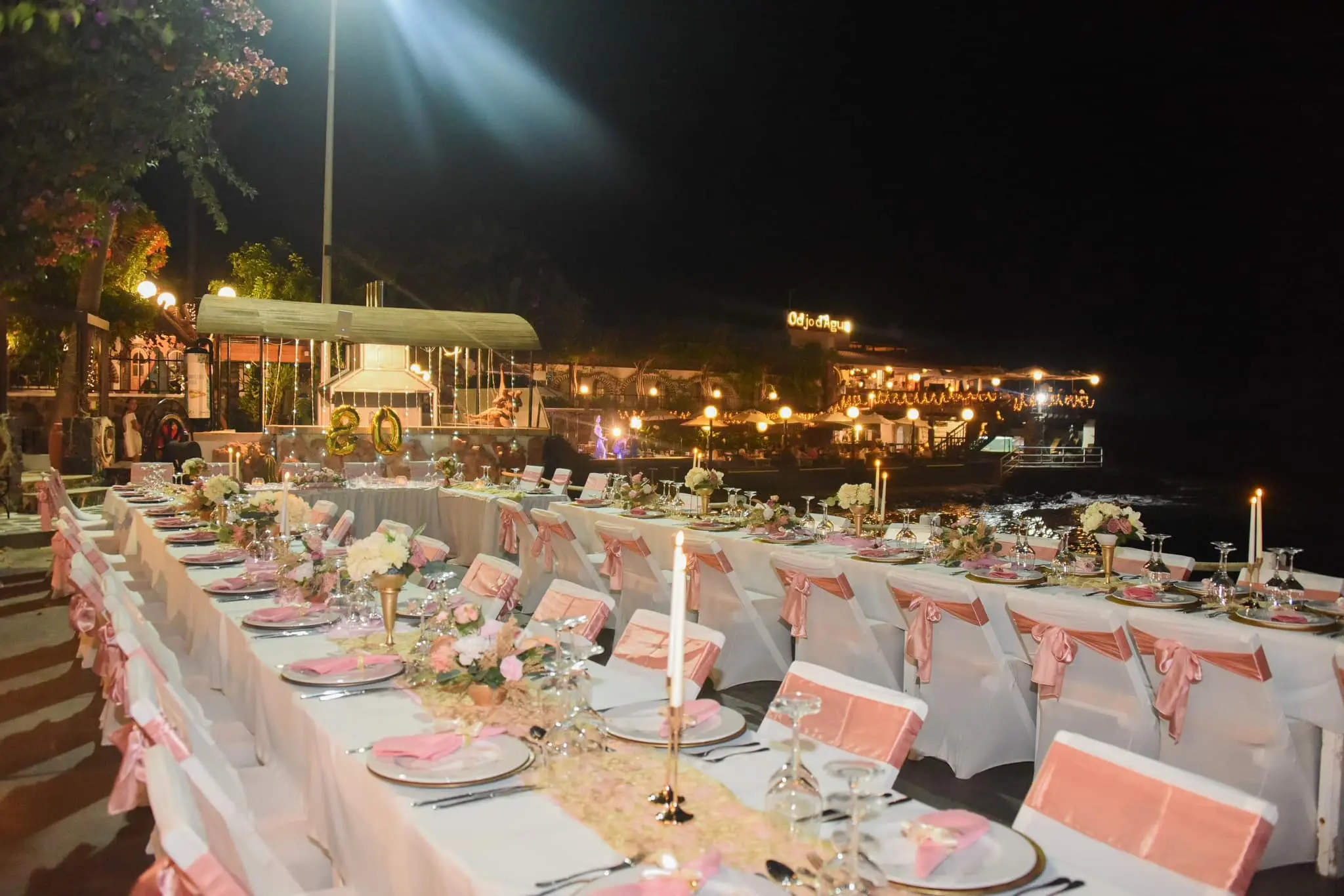 Longa mesa de banquete montada ao ar livre à noite perto do mar, com cadeiras decoradas com fitas cor-de-rosa em Santa Maria, Ilha do Sal, Cabo Verde.