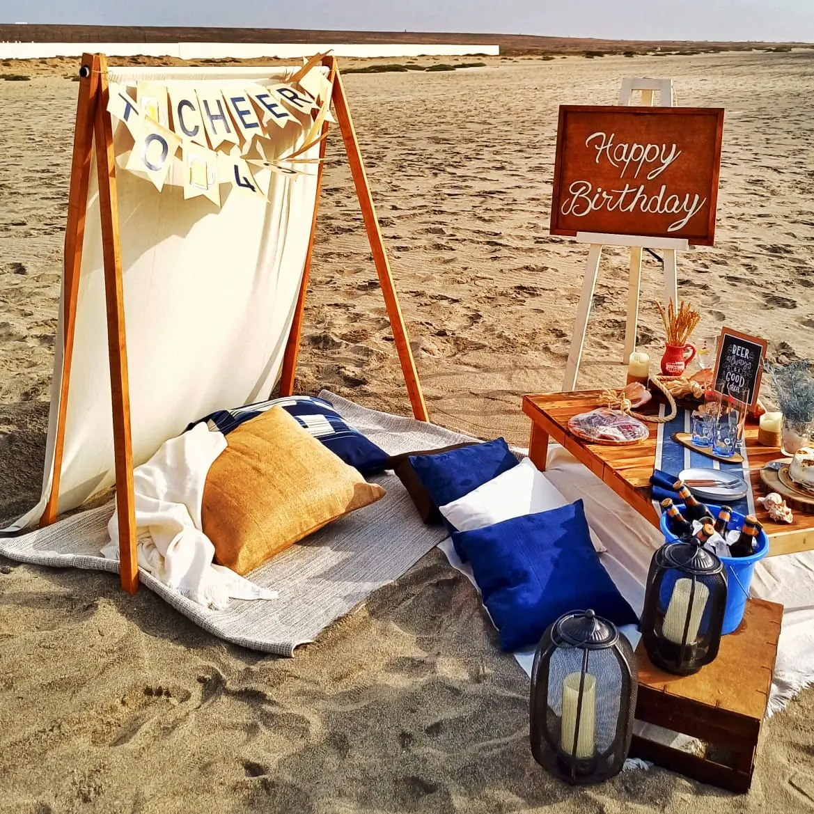 Piquenique de aniversário na praia com tenda decorada, mesa baixa de madeira com bolo, balde de cervejas, almofadas e placa comemorativa. Santa Maria, Ilha do Sal, Cabo Verde.