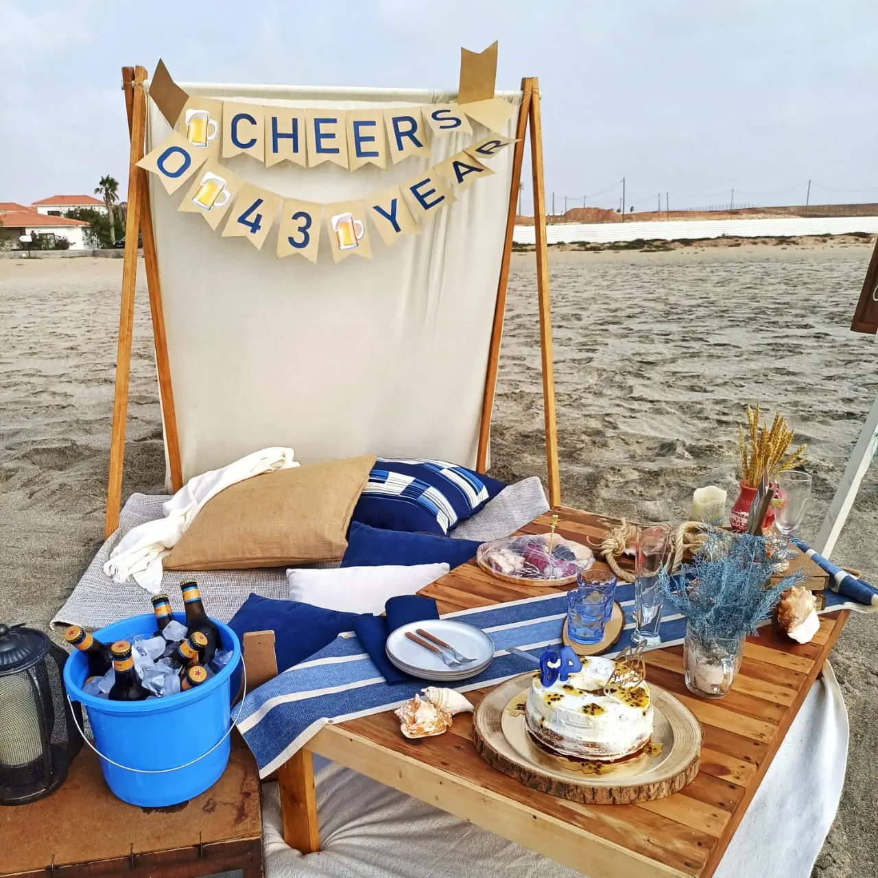 Pormenor de piquenique de 43.º aniversário na praia com bolo de maracujá, pratos azuis, cervejas no gelo e tenda decorada. Santa Maria, Ilha do Sal, Cabo Verde.