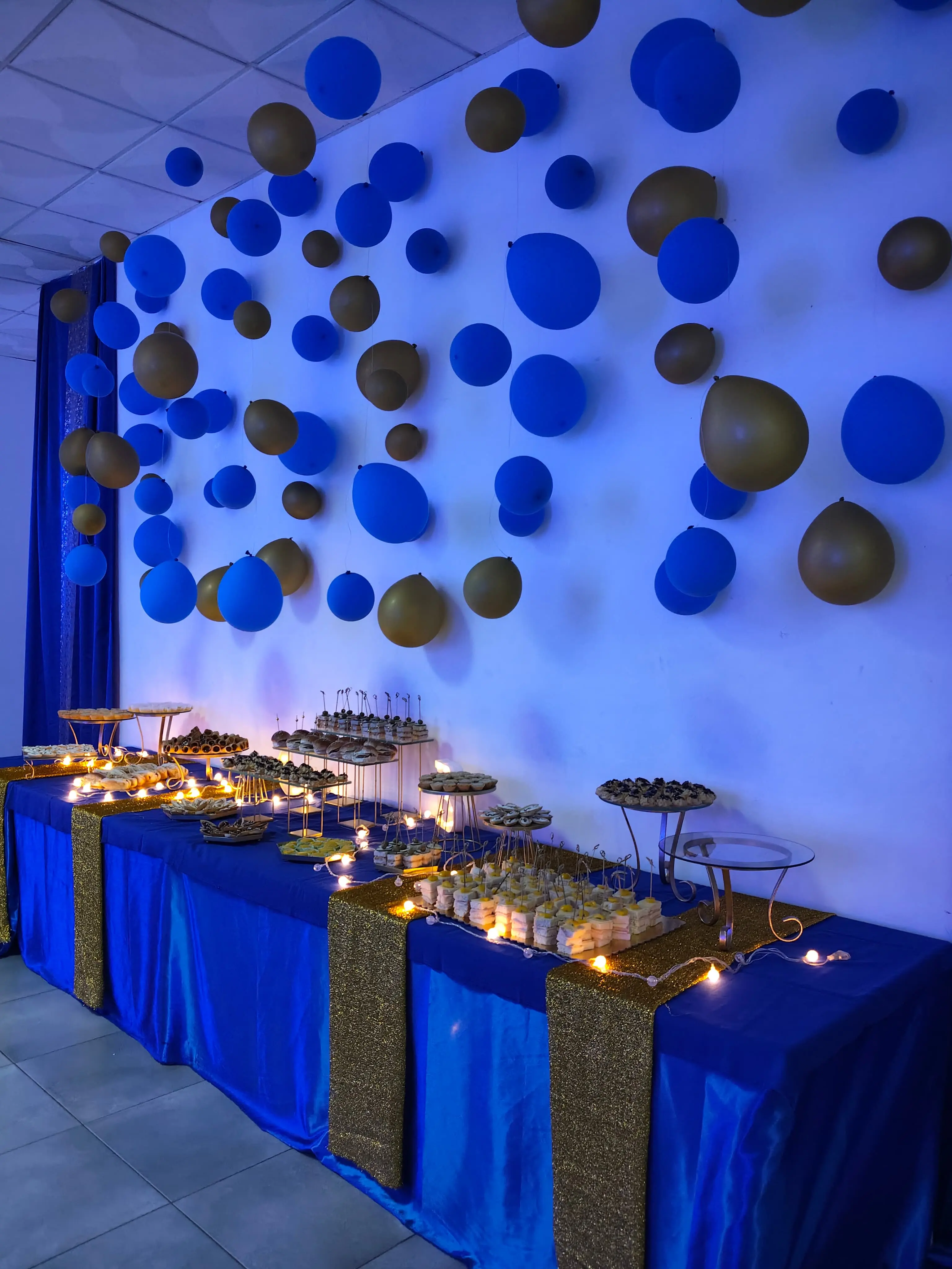 Mesa de buffet decorada com arco de balões azuis e dourados. Toalha azul, passadeiras de lantejoulas douradas e travessas com canapés. Clube ASA, Ilha do Sal, Cabo Verde.