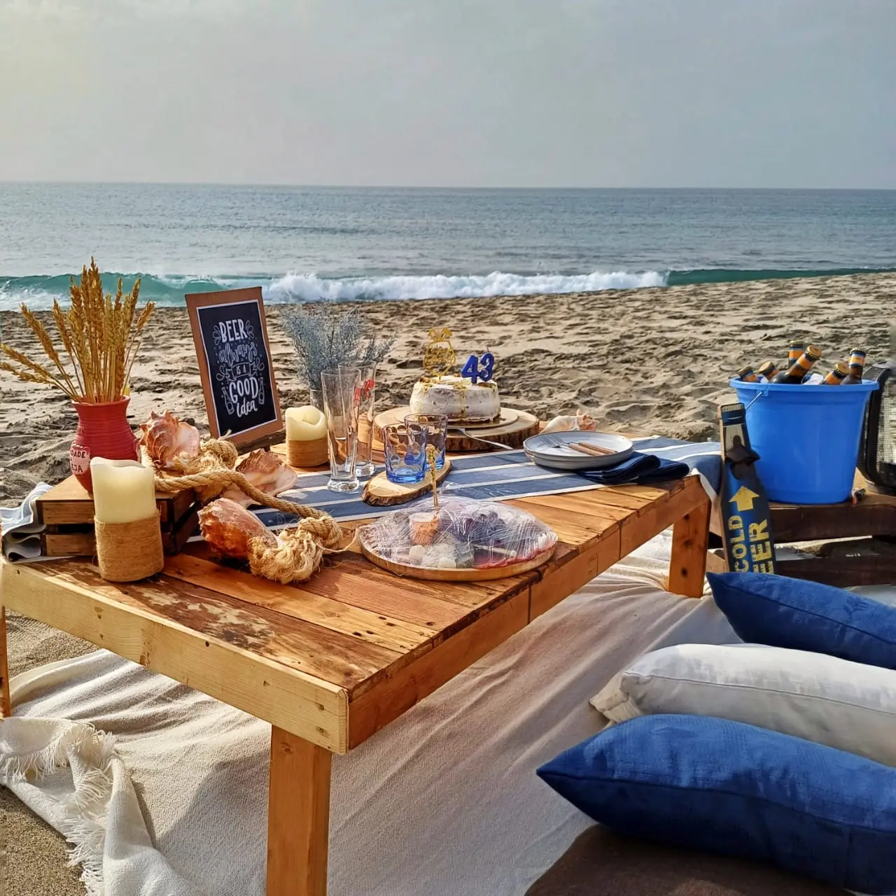 Piquenique ao pôr do sol na praia de Santa Maria com mesa baixa de paletes, bolo, bebidas em balde azul, almofadas confortáveis e placa decorativa. Santa Maria, Ilha do Sal, Cabo Verde.
