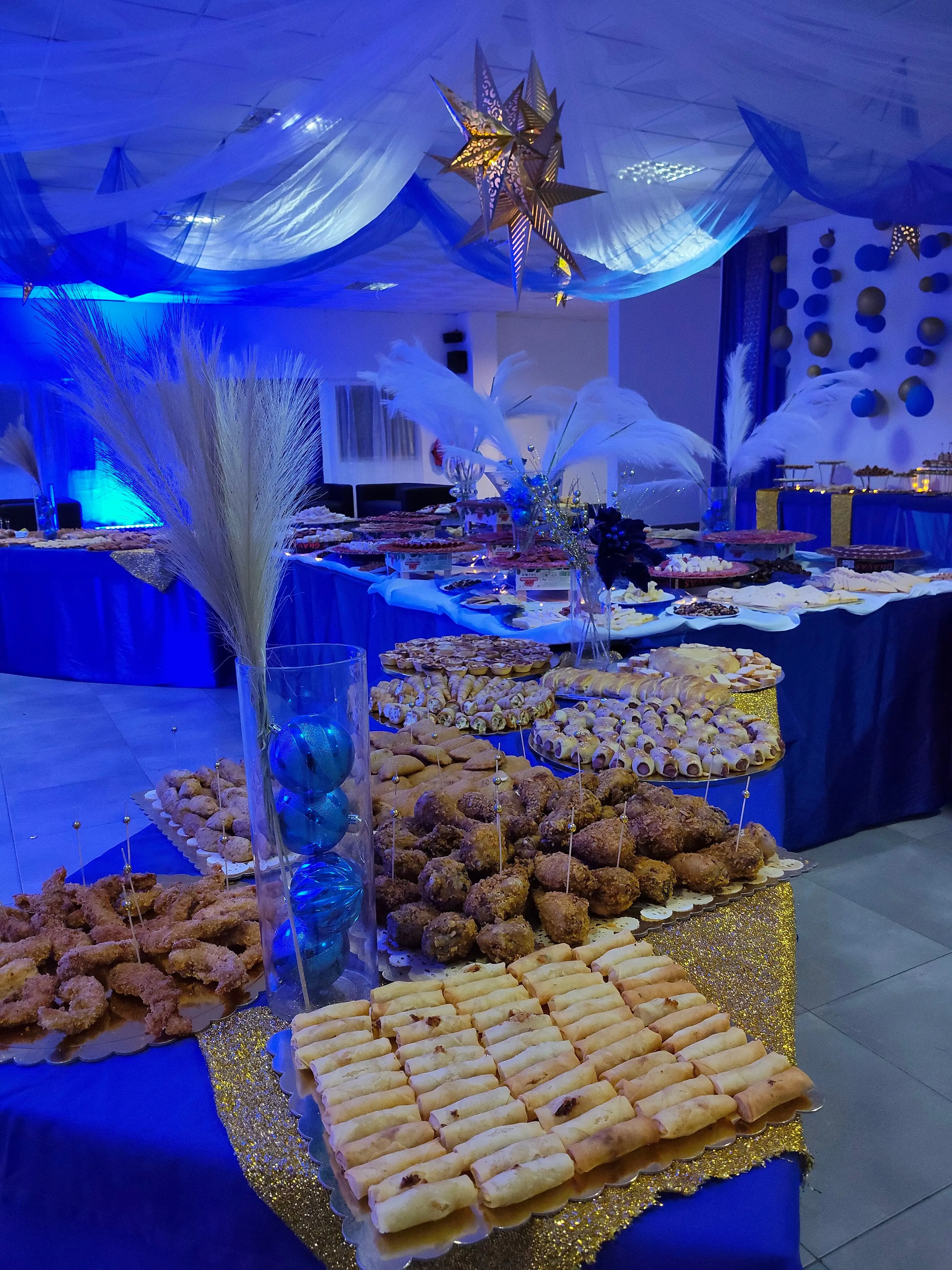 Pormenor de mesa de buffet com toalhas azuis e passadeira de lantejoulas douradas, servindo diversos aperitivos. Decoração com arranjos rústicos no Clube ASA, Ilha do Sal, Cabo Verde.