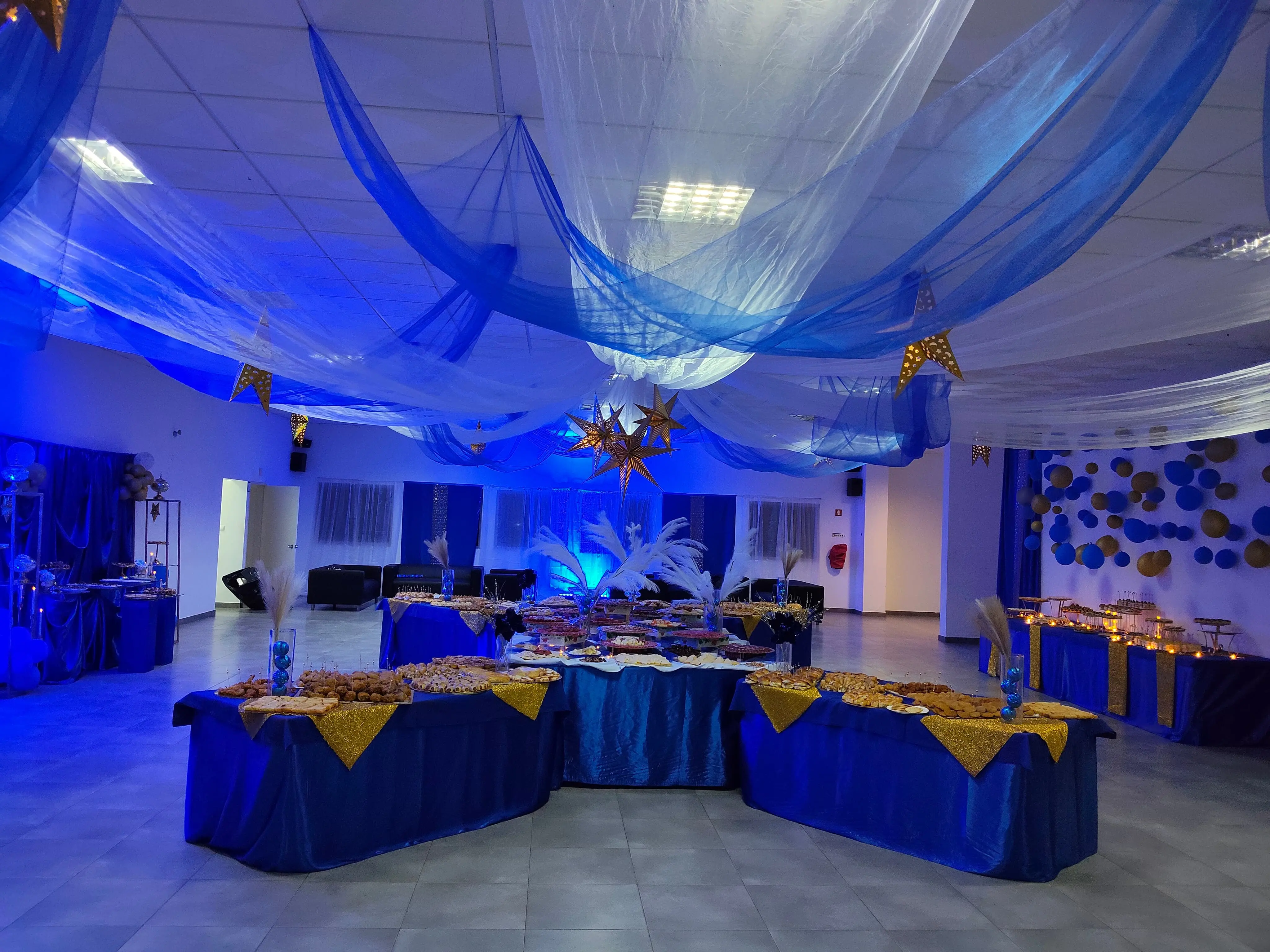 Salão de festas decorado com tecidos azuis e brancos no teto, estrelas douradas suspensas e mesas de buffet com toalhas azuis. Clube ASA, Ilha do Sal, Cabo Verde.