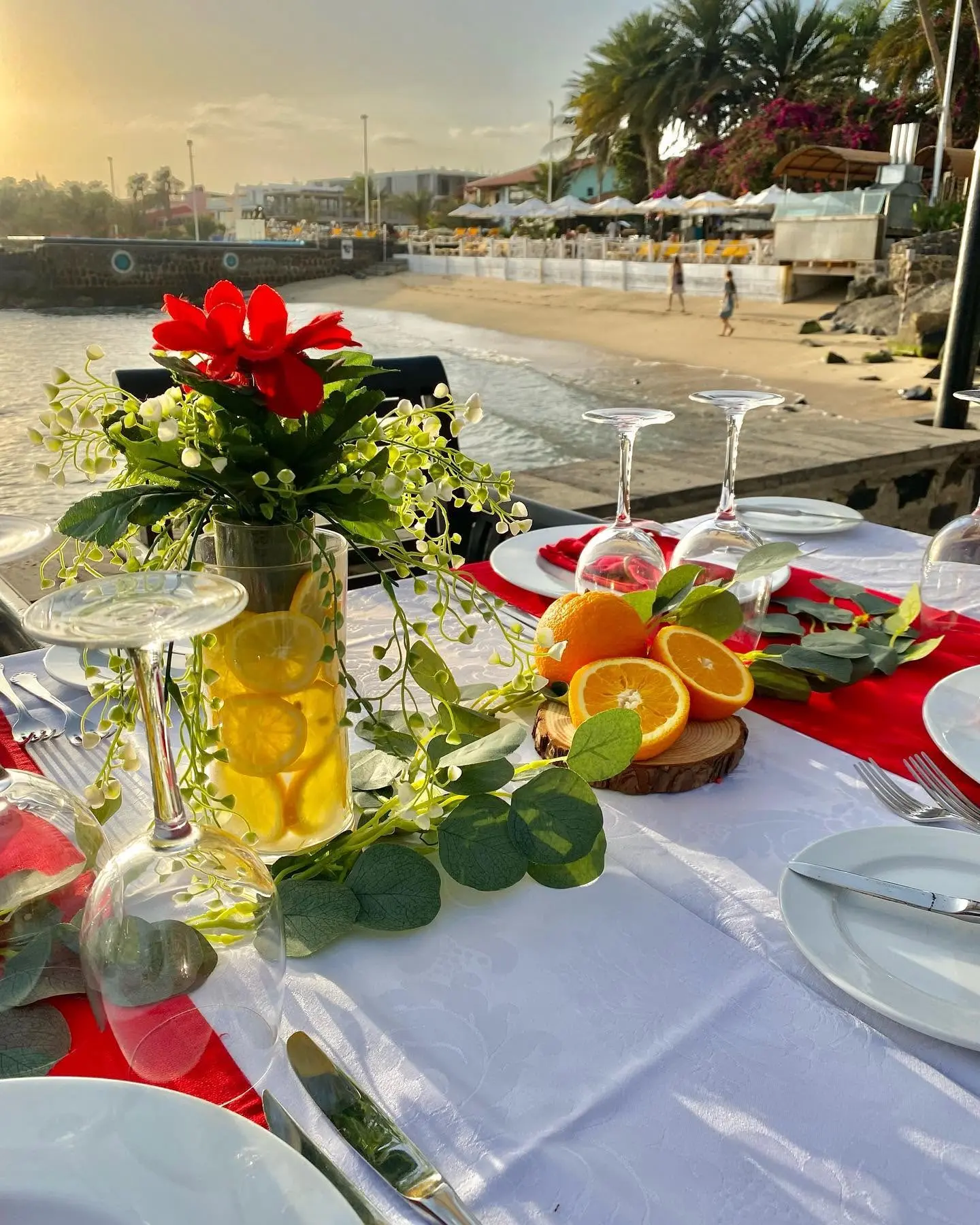 Centro de mesa com vaso de vidro com rodelas de limão, folhagens e flores vermelhas. Laranjas frescas sobre bases de madeira. Santa Maria, Ilha do Sal, Cabo Verde.