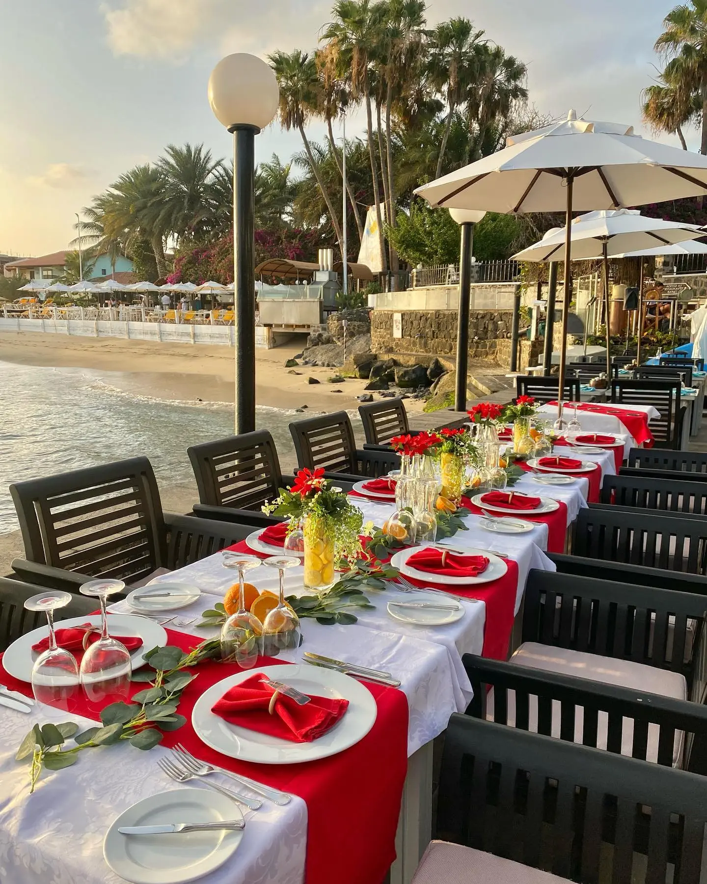 Mesa comprida montada para refeição à beira-mar ao pôr do sol, com toalha branca, passadeira vermelha e arranjos florais com limões e laranjas. Santa Maria, Ilha do Sal, Cabo Verde.