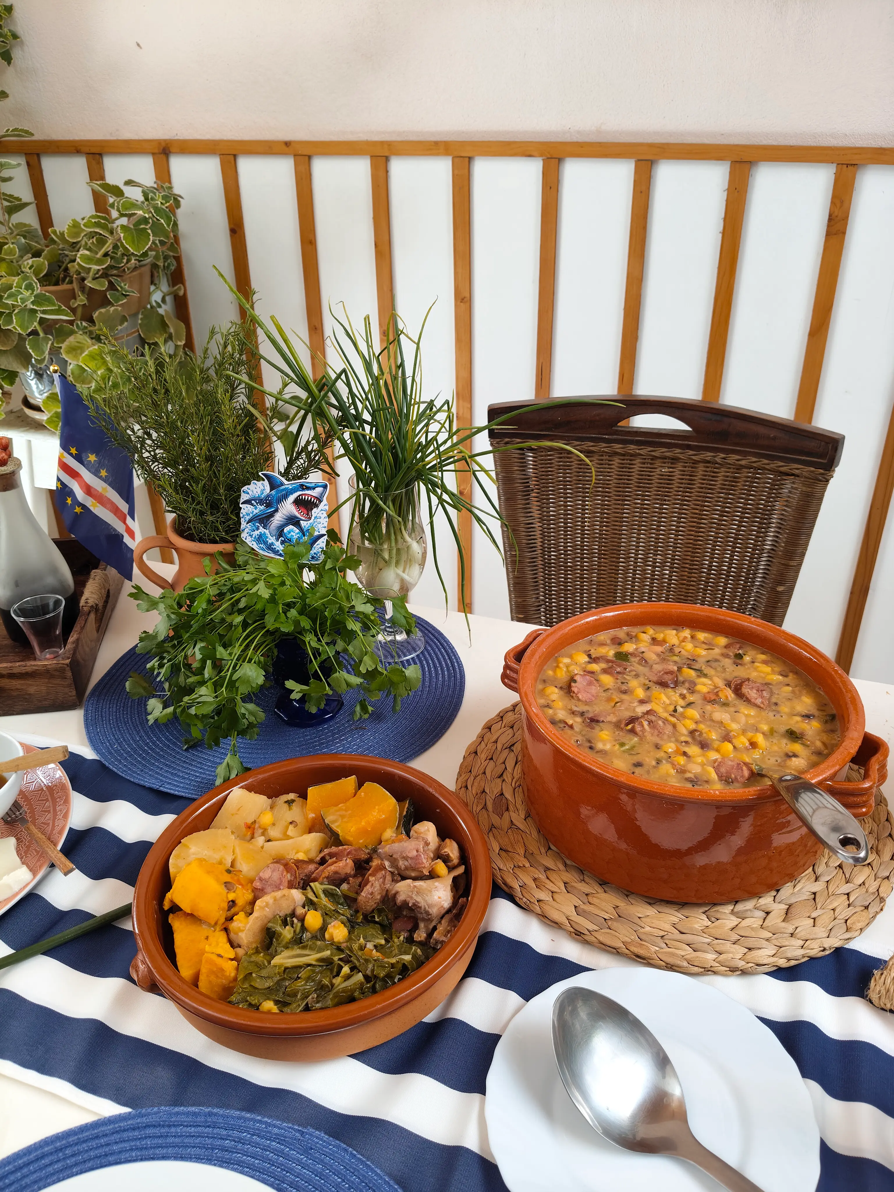 Mesa farta com comida tradicional, incluindo panela de barro com cachupa, legumes e carne cozidos, e uma pequena bandeira de Cabo Verde. Ilha do Sal, Cabo Verde.