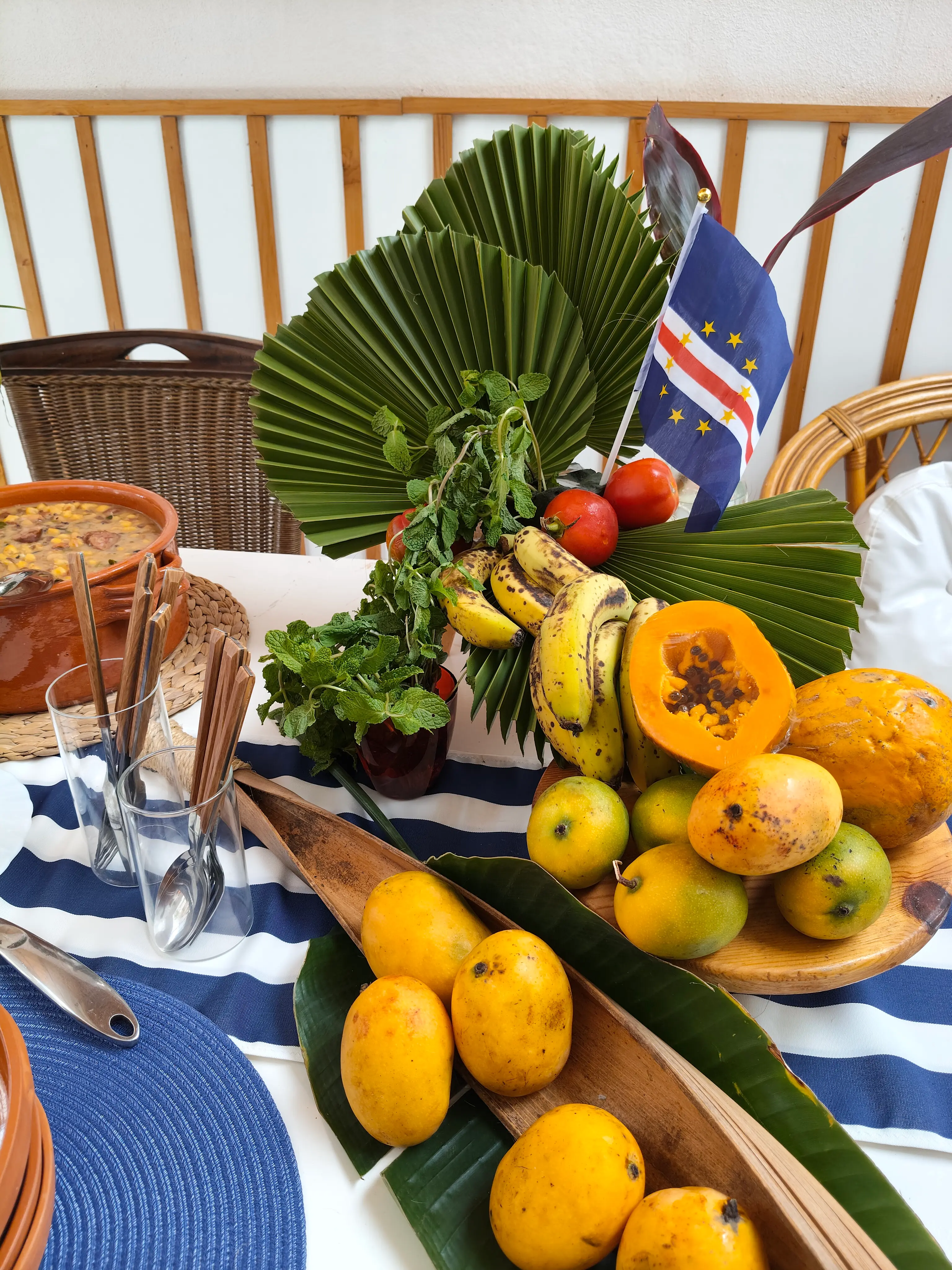 Arranjo de frutas tropicais com mamão, mangas, bananas e tomates sobre travessa de madeira e cesta de palha, decorado com a bandeira de Cabo Verde. Ilha do Sal, Cabo Verde.