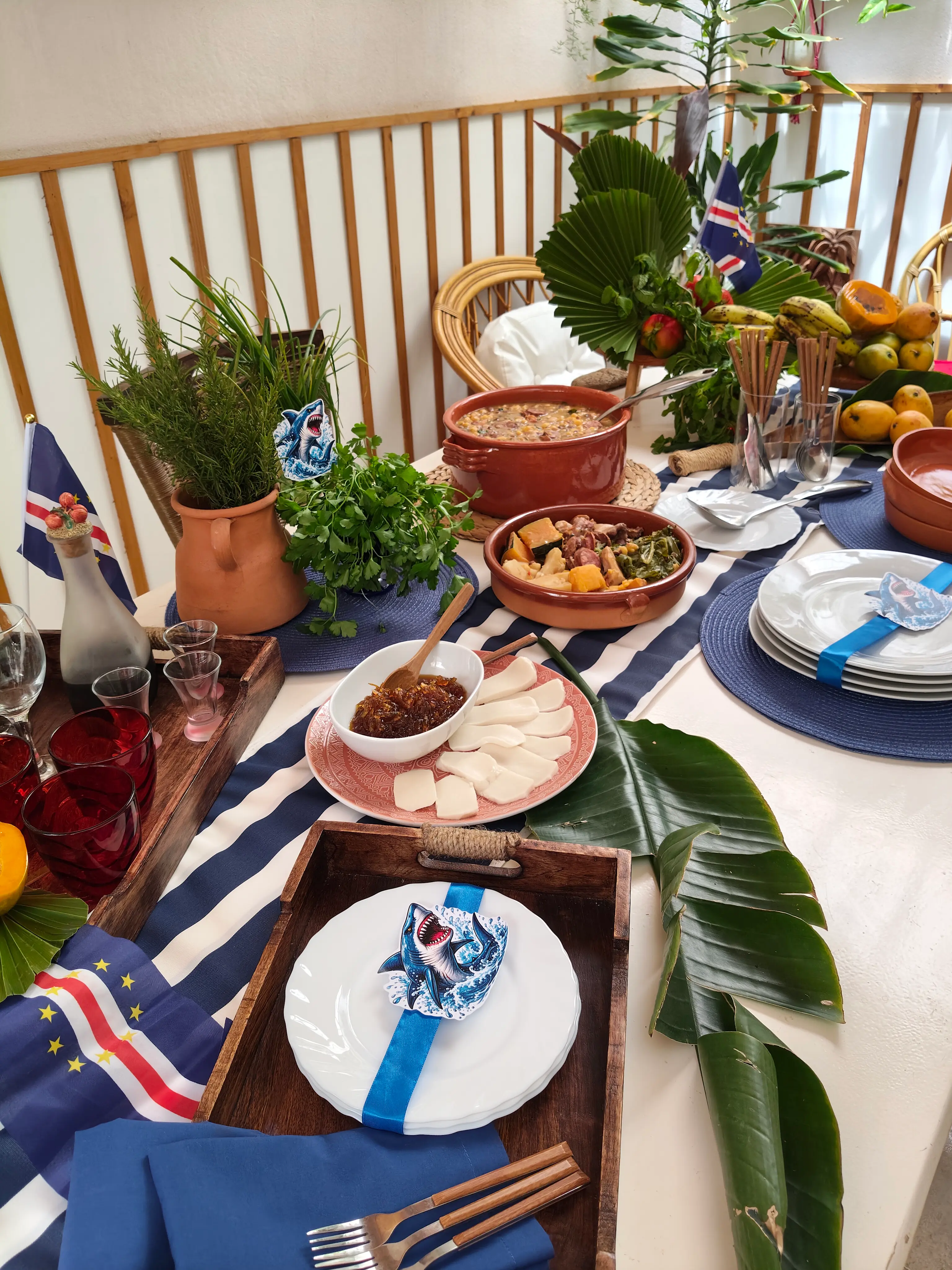 Mesa com pratos típicos de Cabo Verde, incluindo cachupa em panela de barro, queijo tradicional com doce de papaia, porta-guardanapo de tubarão e a bandeira nacional. Ilha do Sal, Cabo Verde.