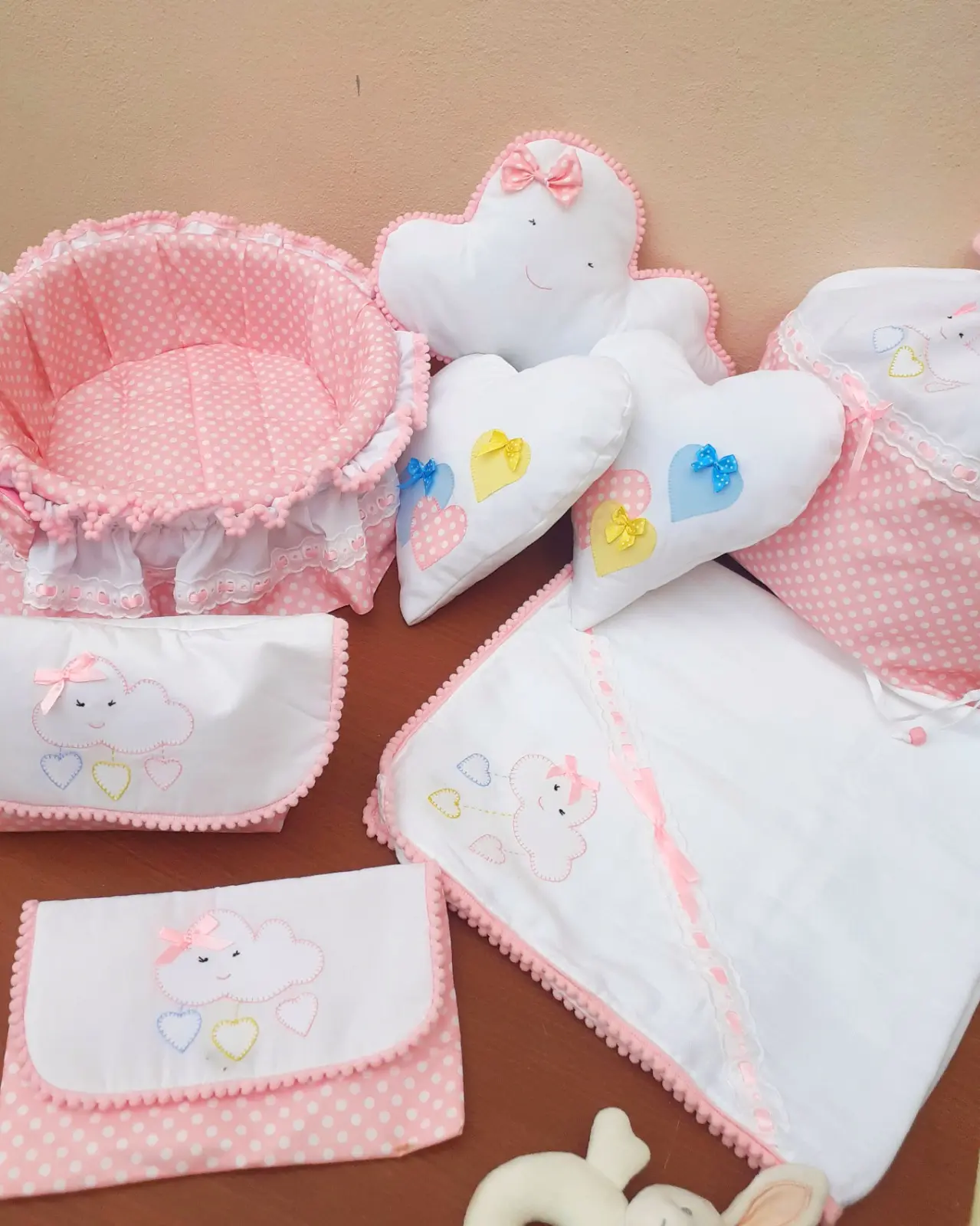 Conjunto de bebé cor-de-rosa com padrão de bolinhas brancas, composto por cesto acolchoado, almofadas de nuvem e coração, e bolsas. Ilha do Sal, Cabo Verde.