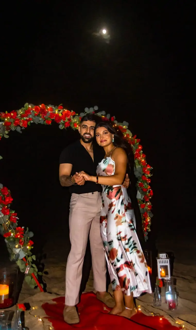 Casal recém-noivo sorridente e de mãos dadas à noite na praia de Santa Maria, em frente a um grande arco em forma de coração de rosas vermelhas iluminadas. Ilha do Sal, Cabo Verde.
