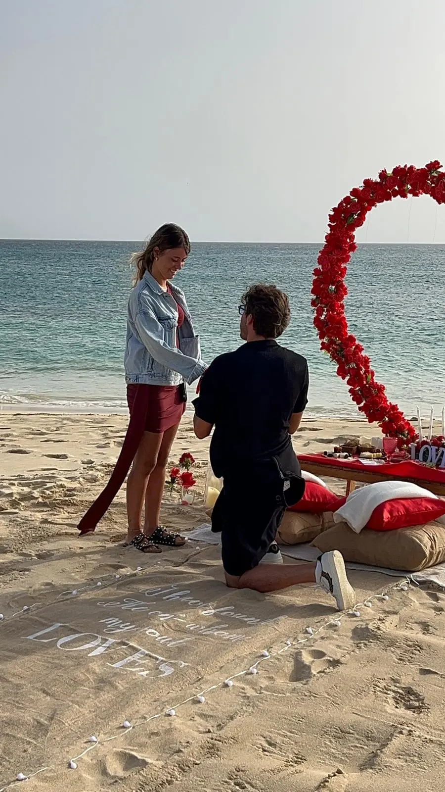 Pedido de casamento com piquenique na praia de Santa Maria, Ilha do Sal. Homem ajoelhado sobre tapete com a palavra LOVE, segurando a aliança para a noiva, com coração de rosas vermelhas ao fundo.