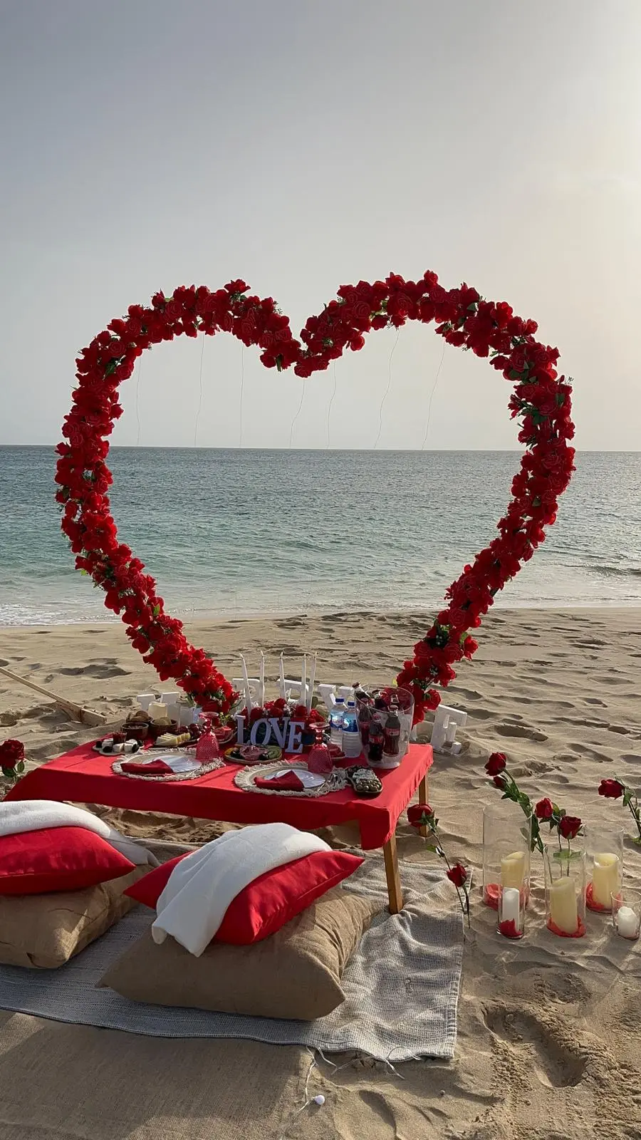 Vista detalhada do cenário romântico na praia de Santa Maria, Ilha do Sal, com coração de rosas vermelhas, mesa com letreiro LOVE, velas e almofadas vermelhas na areia.
