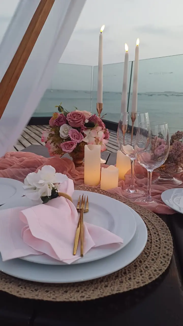 Detalhe de mesa de jantar romântica com guardanapos cor-de-rosa, argolas de flores brancas, talheres dourados, taças de vinho, arranjo floral rosa e velas altas na praia de Santa Maria, Ilha do Sal, Cabo Verde.