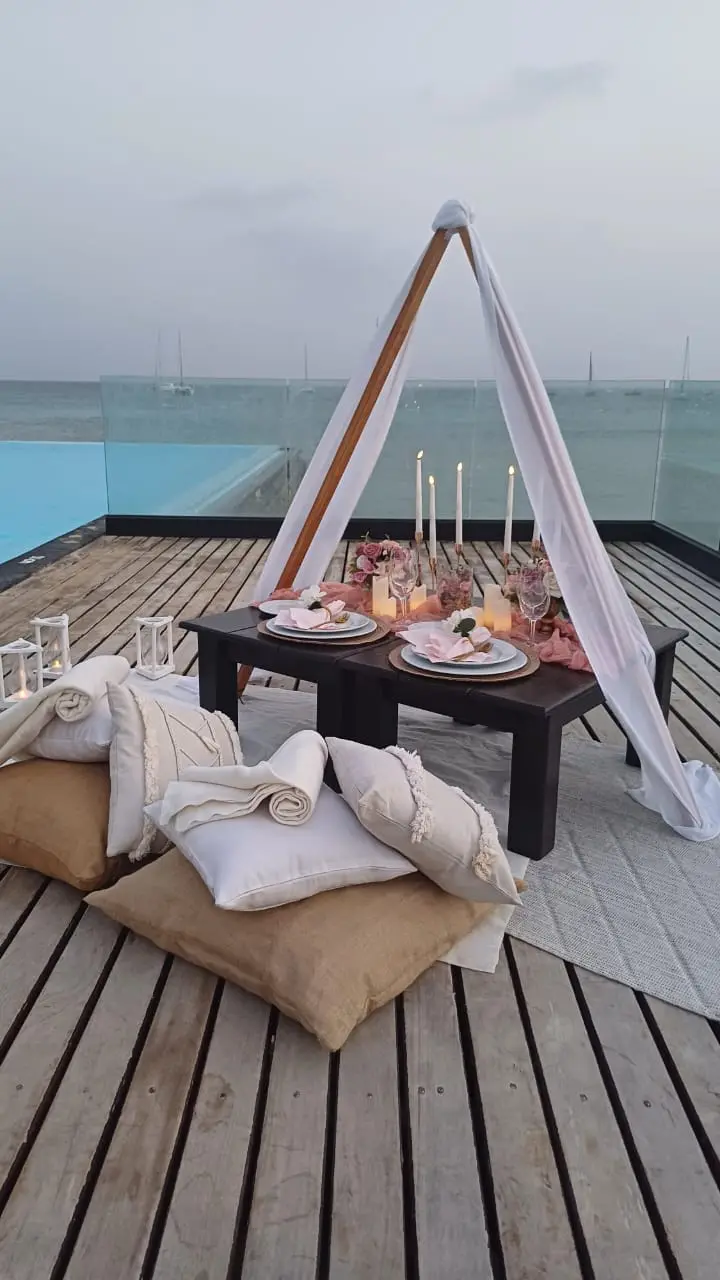 Piquenique romântico em mesa de madeira com vista para o mar na praia de Santa Maria, Ilha do Sal. Tenda branca com tecido transparente, mesa baixa para dois com guardanapos cor-de-rosa, velas, flores e almofadas.