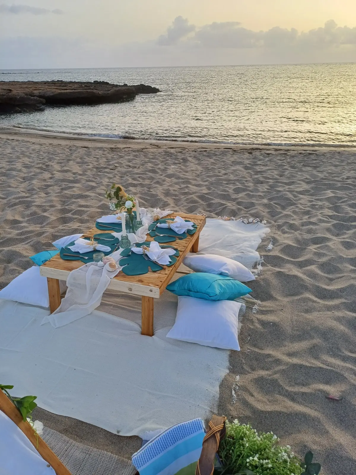 Mesa posta para piquenique com tenda branca, almofadas em tons de azul e um bote tradicional junto à praia da Calheta Funda ao entardecer. Ilha do Sal, Cabo Verde.