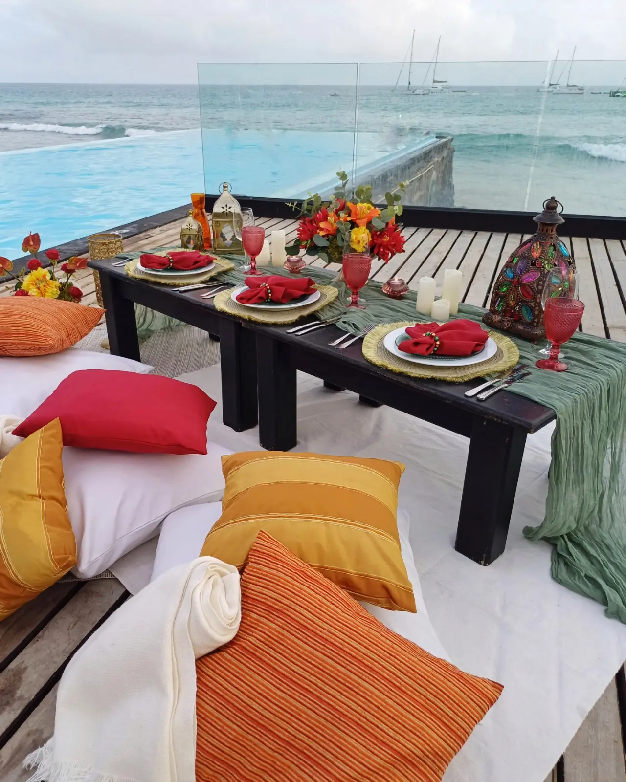 Mesa posta para piquenique com almofadas em tons de laranja, vermelho e amarelo, criando um ambiente acolhedor junto à piscina e ao mar em Santa Maria, Ilha do Sal, Cabo Verde.