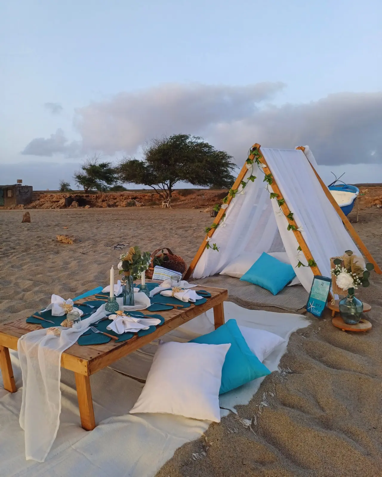 Mesa posta para piquenique com tenda branca e almofadas em tons de azul, com vista para a paisagem rústica junto à praia da Calheta Funda. Ilha do Sal, Cabo Verde.