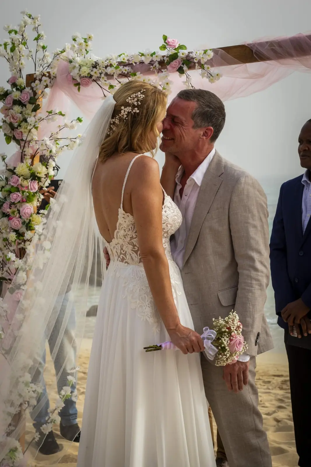 Noivos partilham momento carinhoso e beijo no altar sob arco floral rosa e branco na praia de Santa Maria, Ilha do Sal, Cabo Verde.