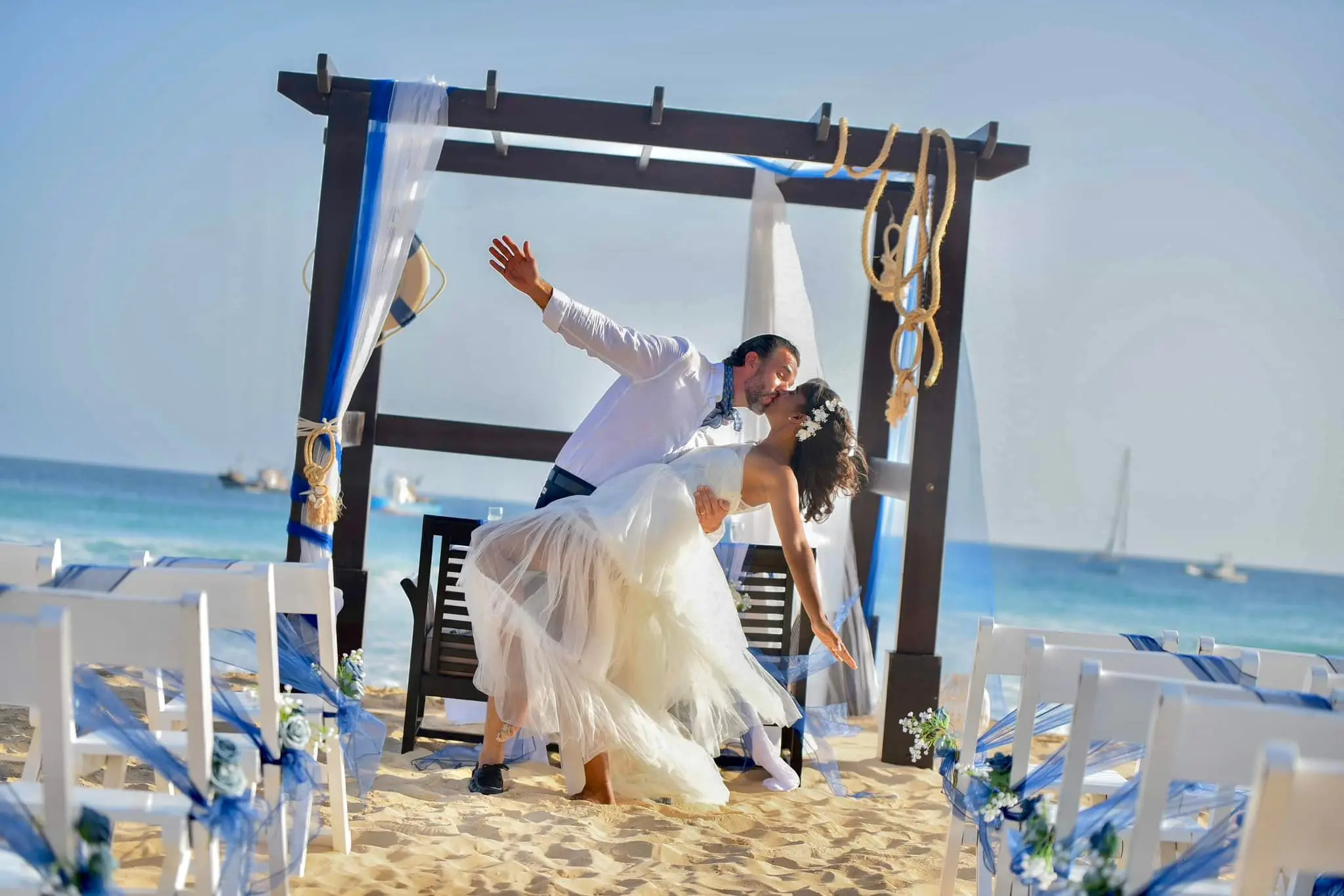 Noivos apaixonados a celebrar o casamento na praia sob uma pérgola decorada em azul e branco. Santa Maria, Ilha do Sal, Cabo Verde.